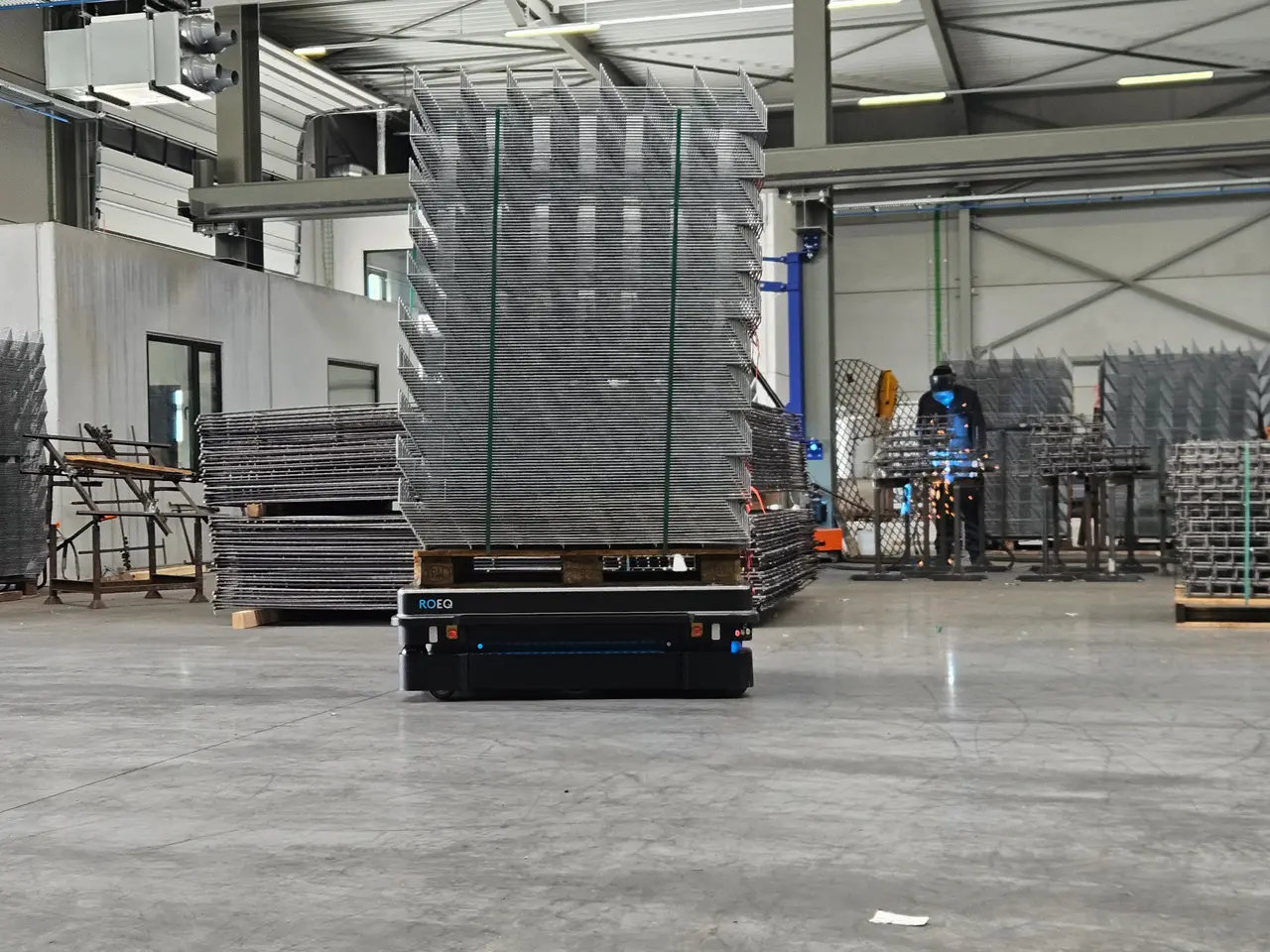 An autonomous mobile robot transporting a tall stack of welded wire-mesh panels across a metalworking floor, a welder at work in the background